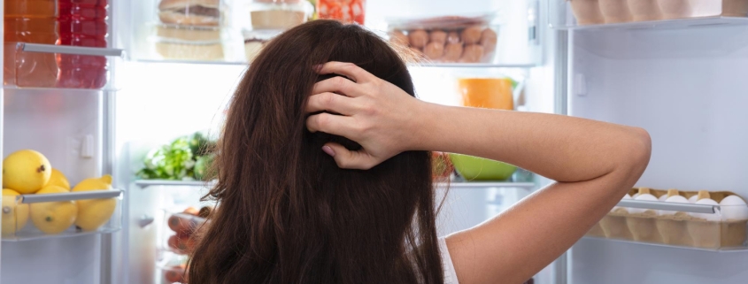 Donna con capelli lunghi che si gratta la testa e guarda in frigo